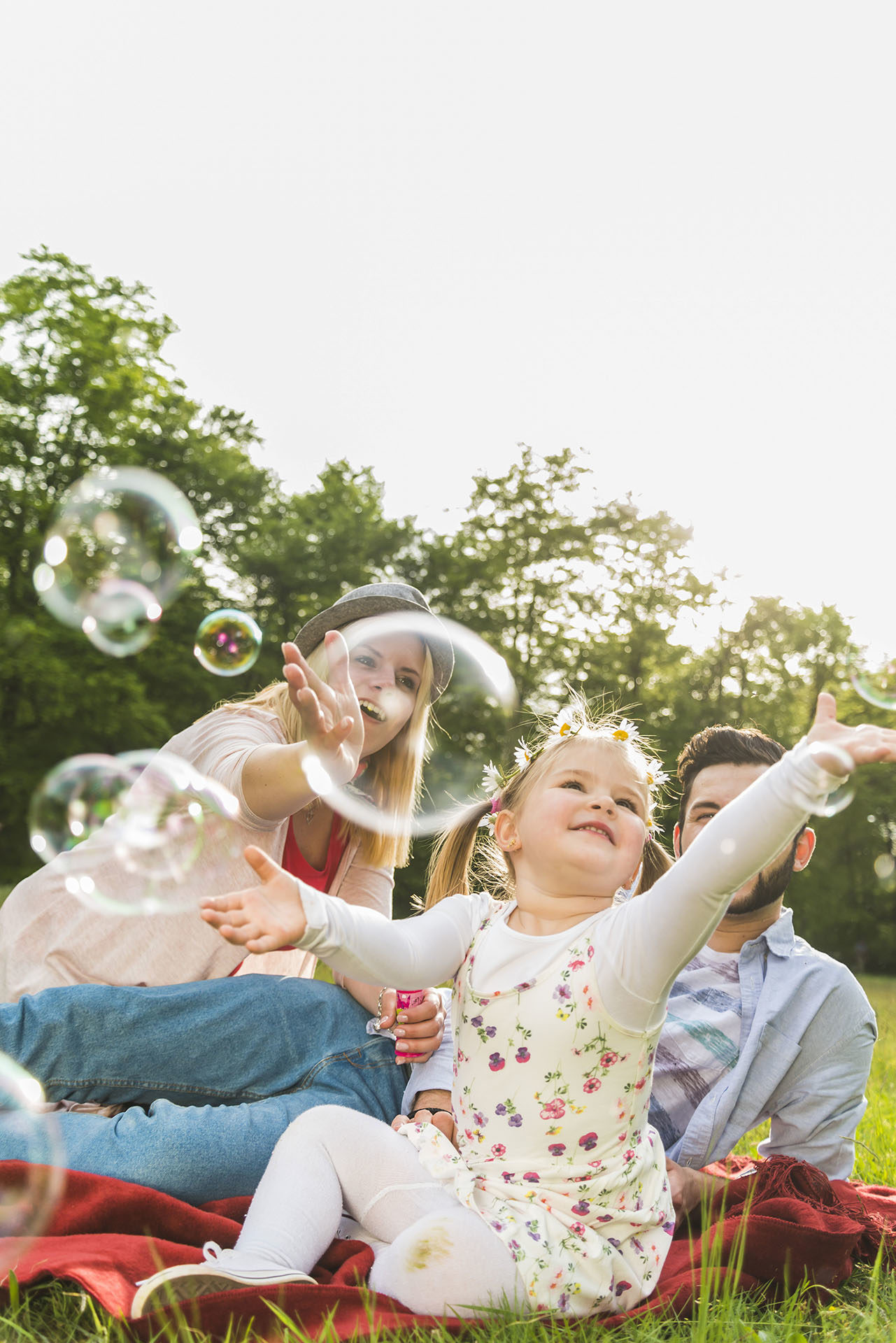Happy family sitting on blanket in meadow catching soap bubbles